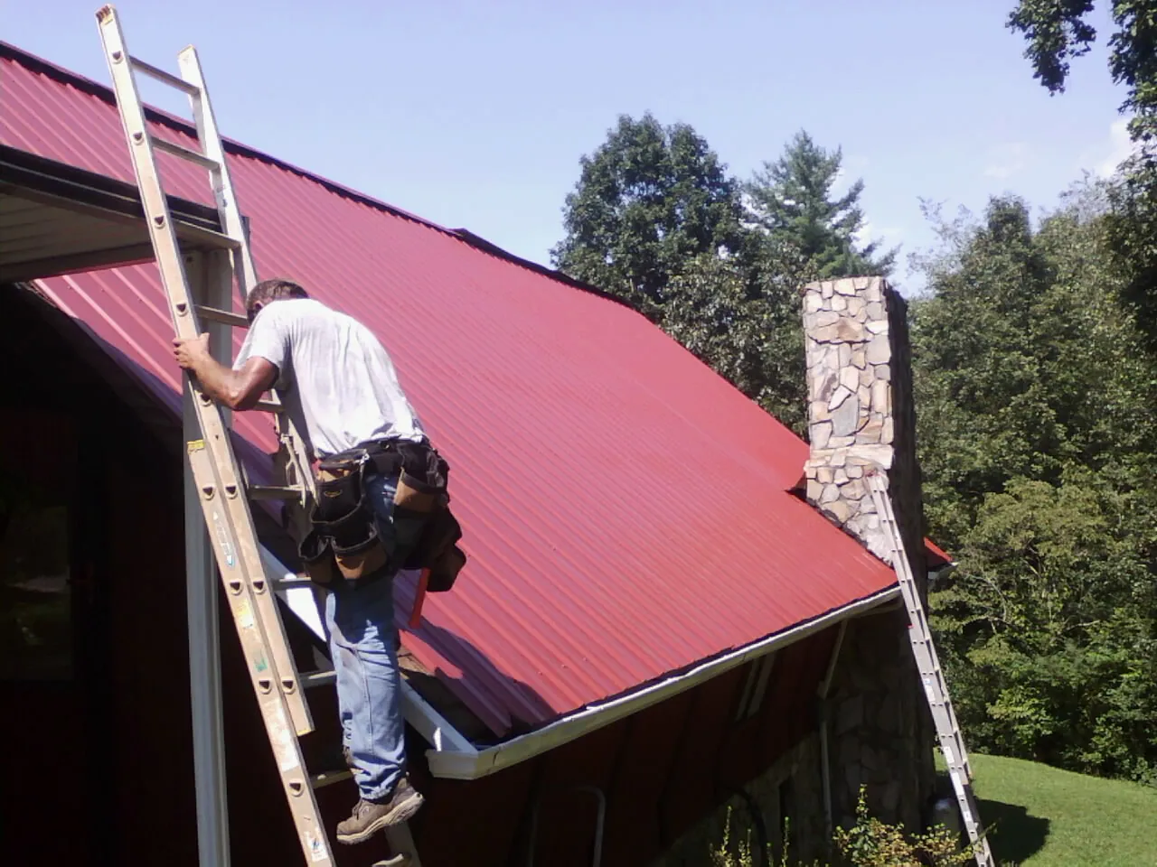 Roofer on a ladder beside a red metal roof and stone chimney — Soffit & Fascia Repair by Roofing Brothers in Gaffney